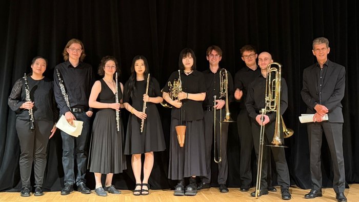 Neun Mitglieder des Ensembles Ribörs mit Klarinetten, Flöten, Horn und Posaunen auf einer Bühne vor schwarzem Vorhang.