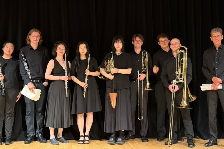 Nine members of the Ribörs Ensemble with clarinets, flutes, horn and trombones on a stage in front of a black curtain.
