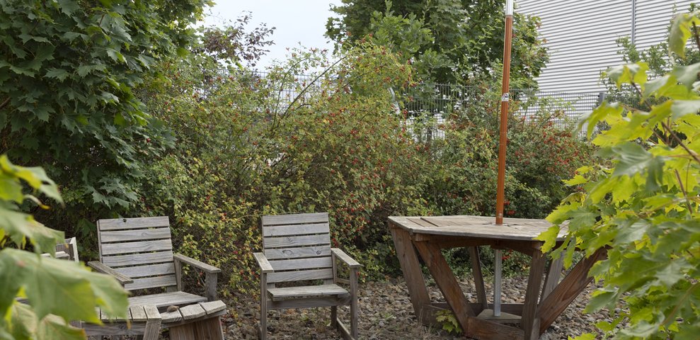 A small garden area with two wooden chairs and a table on a ground covered with pebbles. The area is surrounded by leafy green trees and shrubs. A building with a metal exterior is visible in the background.