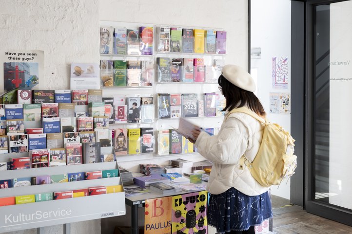 Eine Person in weißer Jacke und Barett steht vor einem Display mit Broschüren in einem Kulturservicezentrum und hält eine Broschüre in der Hand. Das Display enthält Informationen zu Kunst und Veranstaltungen. Helle Plakate und Broschüren sind sichtbar.