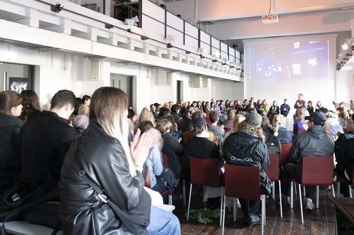 Audience attending a presentation in a spacious, modern room at Speicher XI, part of the University of the Arts Bremen. The setting features high ceilings and a large projection screen.