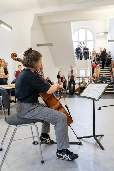 Cellist:in spielt in einem Flur des Fachbereichs Musik der HfK Bremen, während im Hintergrund das Publikum auf Treppen sitzt. Heller Raum mit natürlichem Licht durch große Fenster.