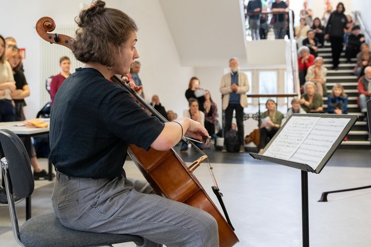 Cellist performing in a hallway at the HfK Bremen music department, with an audience seated on stairs in the background. Bright space with natural light from large windows.