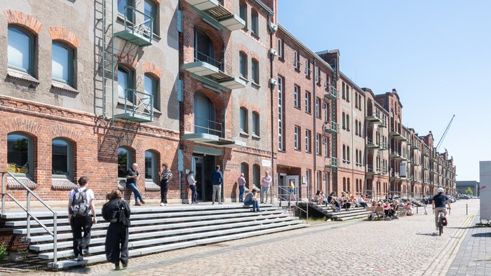 Exterior view of Speicher XI at HfK Bremen, showcasing historic brick architecture with students and visitors interacting on steps and pathways, under a clear blue sky.