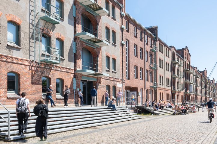 Exterior view of Speicher XI at HfK Bremen, showcasing historic brick architecture with students and visitors interacting on steps and pathways, under a clear blue sky.