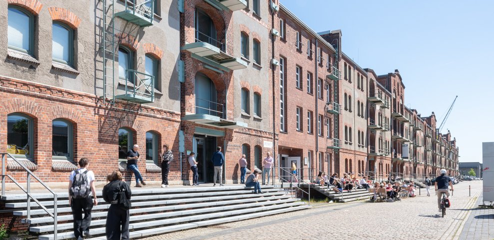 Außenansicht des Speicher XI an der Hochschule für Künste Bremen, die historische Backsteinarchitektur zeigt. Studierende und Besucher:innen interagieren auf den Stufen und Wegen unter einem klaren blauen Himmel.