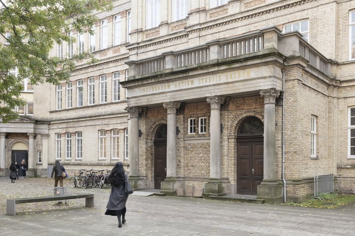 Historisches Gebäude an der Dechanatstraße, in dem der Fachbereich Musik der Hochschule für Künste Bremen untergebracht ist. Es zeigt klassische Architektur mit Säulen und großen Fenstern. Vor dem Gebäude sind Studierende versammelt, und in der Nähe sind Fahrräder abgestellt.