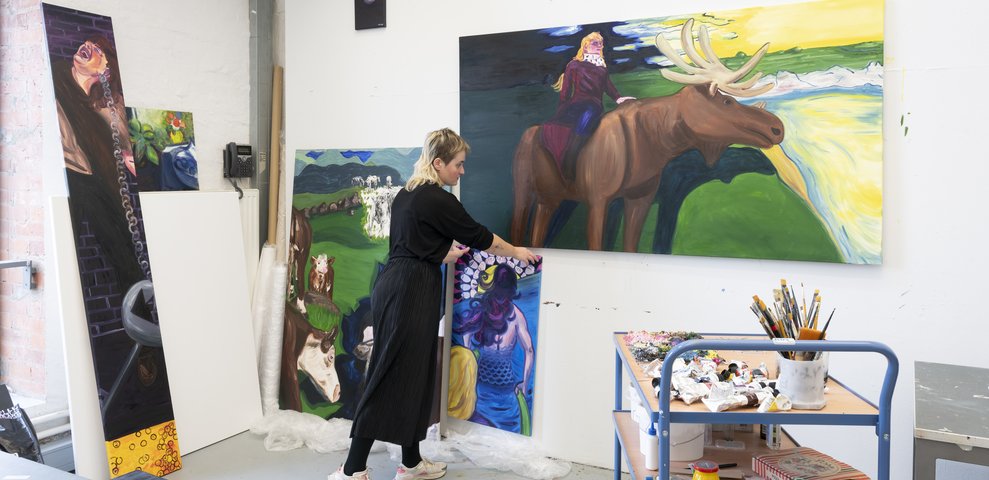 Student:in arrangiert großformatige figurative Gemälde in einem Atelier.