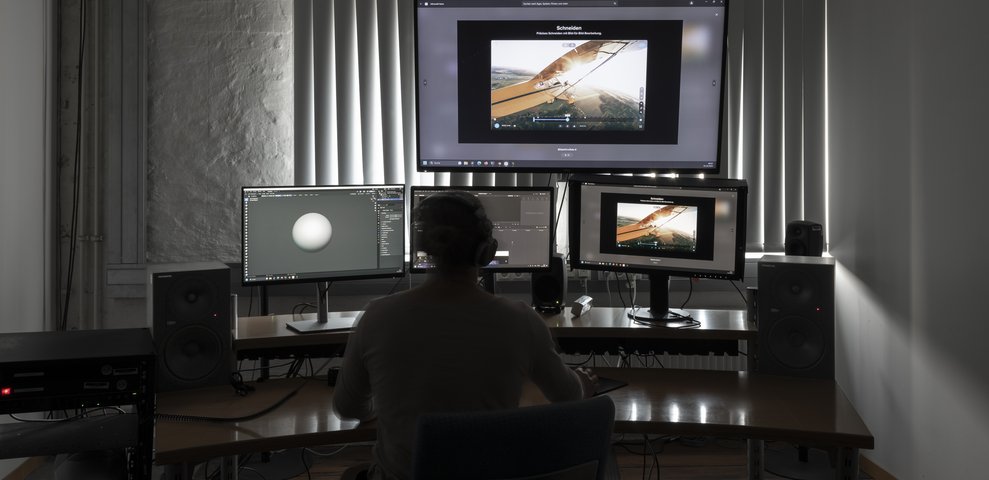 Film editing studio with multiple monitors and audio speakers, featuring video editing software. A person wearing headphones works at a desk. Part of the Werkstätte und Studios at HfK Bremen.