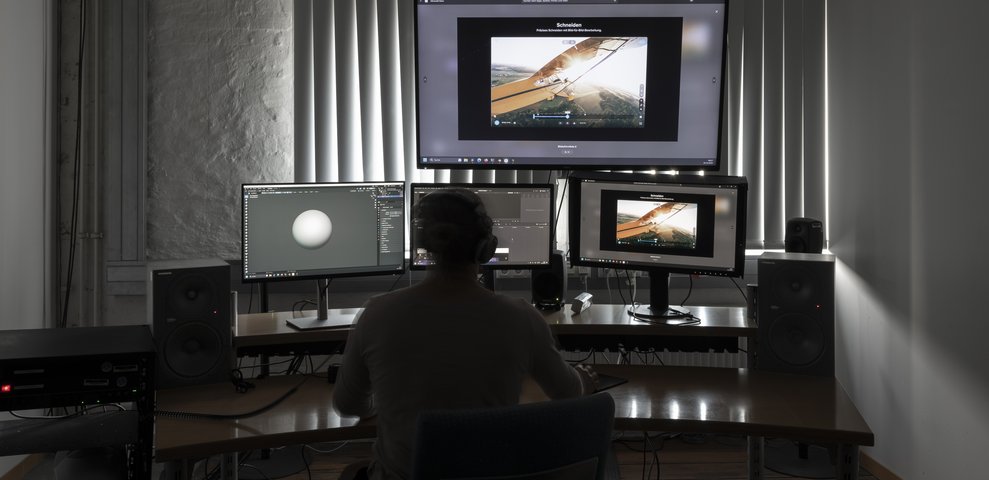 Film editing studio with multiple monitors and audio speakers, featuring video editing software. A person wearing headphones works at a desk. Part of the Werkstätte und Studios at HfK Bremen.