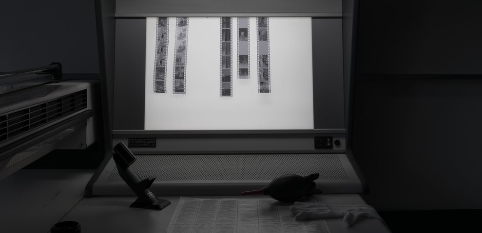 Light table in HfK Bremen photography studio displaying film strips, with a magnifier, gloves, and cleaning blower nearby.