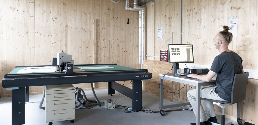 Person working at a computer station in a wood-paneled HfK Bremen studio, next to a large graphics cutter on a table.
