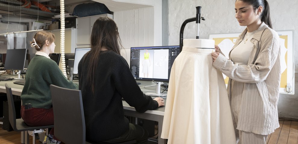 Studierende arbeiten im Mode-CAD-Studio der HfK Bremen und nutzen Computer sowie eine Schneiderpuppe für die Gestaltung von Kleidung.