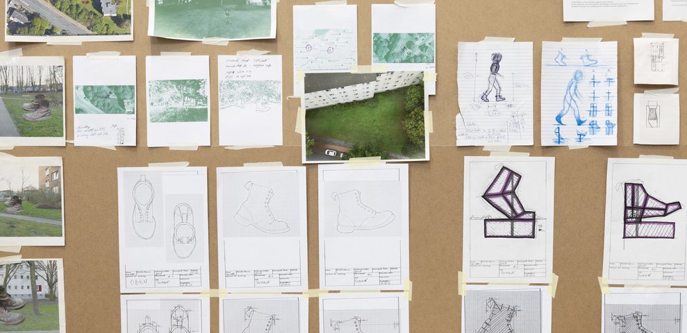 Skizzen und Fotos von Stiefeln, Landschaftsaufnahmen und Notizen sind an eine Korkwand geheftet, um ein Projekt oder eine Installation zu planen.
