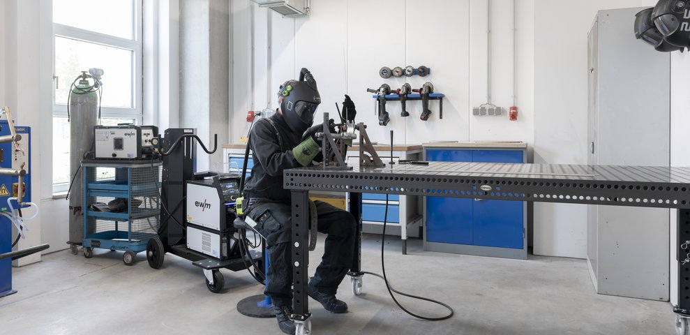 Person welding at a workstation in the Metal Workshop of HfK Bremen, wearing protective gear and surrounded by tools and equipment.