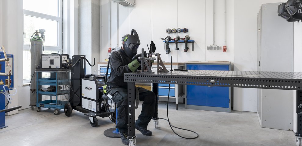 Person welding at a workstation in the Metal Workshop of HfK Bremen, wearing protective gear and surrounded by tools and equipment.