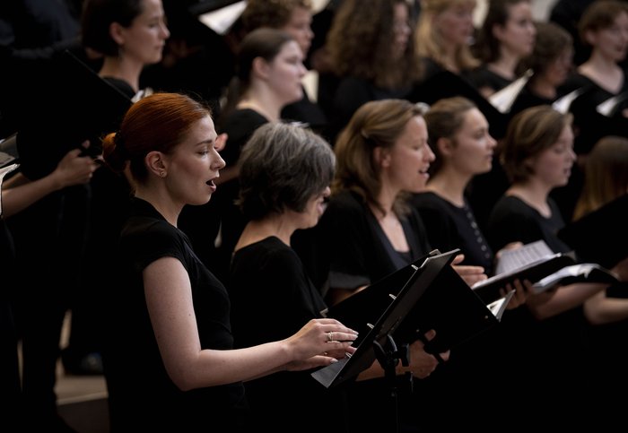 Weibliche Chormitglieder singen, halten Notenblätter, mit Fokus auf eine rothaarige Sängerin im Vordergrund.