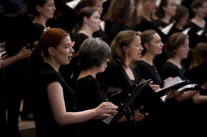 Female choir members singing, holding sheet music, with a focus on a red-haired singer in the foreground.