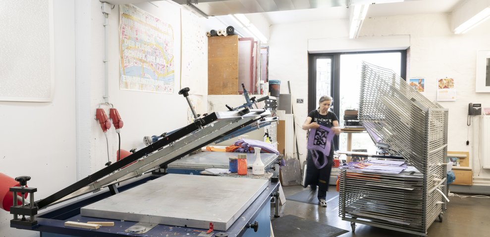 Siebdruckwerkstatt an der HfK Bremen mit Geräten und Trockengestellen, eine Person arbeitet an einem Textilprojekt. Wände zeigen bunte Drucke und Werkzeuge sind im Arbeitsbereich organisiert.