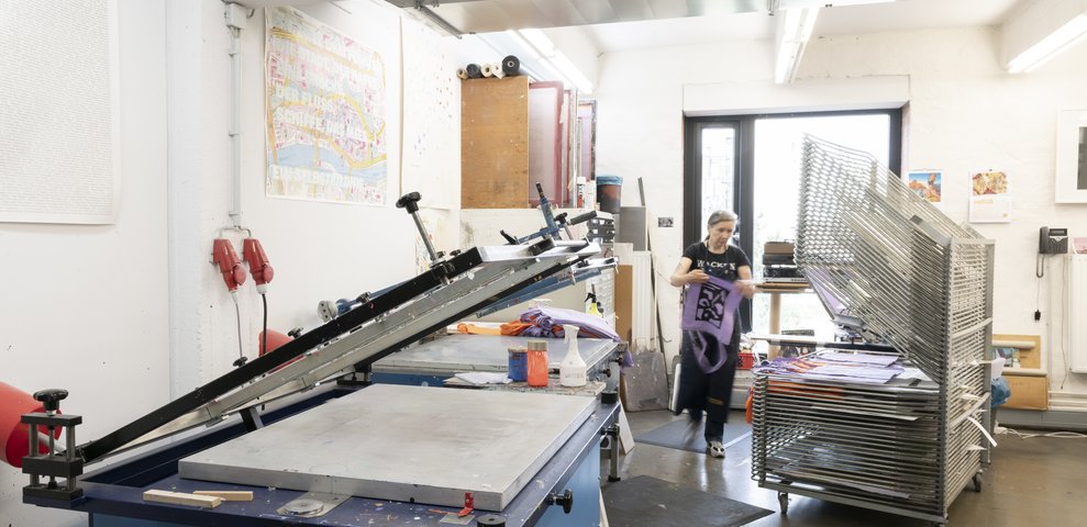 Siebdruckwerkstatt an der HfK Bremen mit Geräten und Trockengestellen, eine Person arbeitet an einem Textilprojekt. Wände zeigen bunte Drucke und Werkzeuge sind im Arbeitsbereich organisiert.