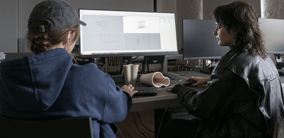 Zwei Studierende arbeiten in der 3D-Werkstatt der HfK Bremen an 3D-Design-Software an Computern, mit 3D-gedruckten Modellen auf dem Tisch.