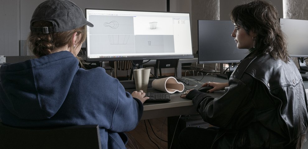Zwei Studierende arbeiten in der 3D-Werkstatt der HfK Bremen an 3D-Design-Software an Computern, mit 3D-gedruckten Modellen auf dem Tisch.