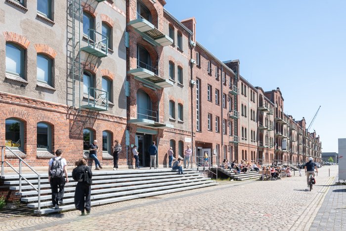 Historic brick facade of Speicher XI at HfK Bremen with people socializing on steps and a cyclist passing by on a sunny day.