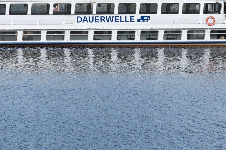 Das Veranstaltungsschiff MS Dauerwelle der Hochschule für Künste Bremen auf dem Wasser mit Spiegelung, der Name der Hochschule ist an der Seite zu sehen.