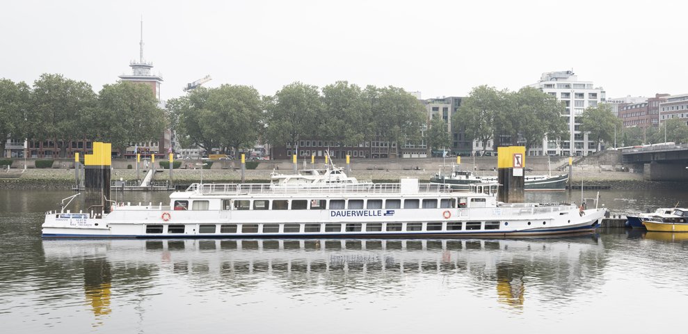 Das Ausstellungsschiff MS Dauerwelle der Hochschule für Künste Bremen liegt an einem Ufer vor Anker. Im Hintergrund sind städtische Gebäude und Bäume zu sehen.