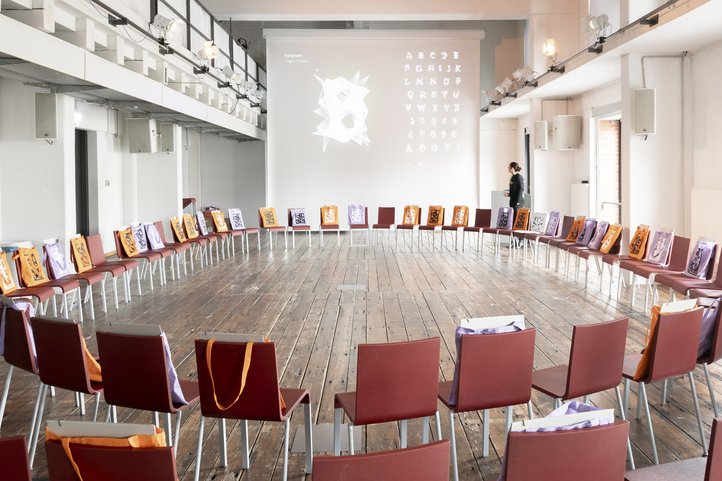 Auditorium im Speicher XI der HfK Bremen mit stuhlkreisförmig angeordneten Stühlen, auf denen jeweils Taschen liegen, und einer projizierten Typografieanzeige an der Wand.