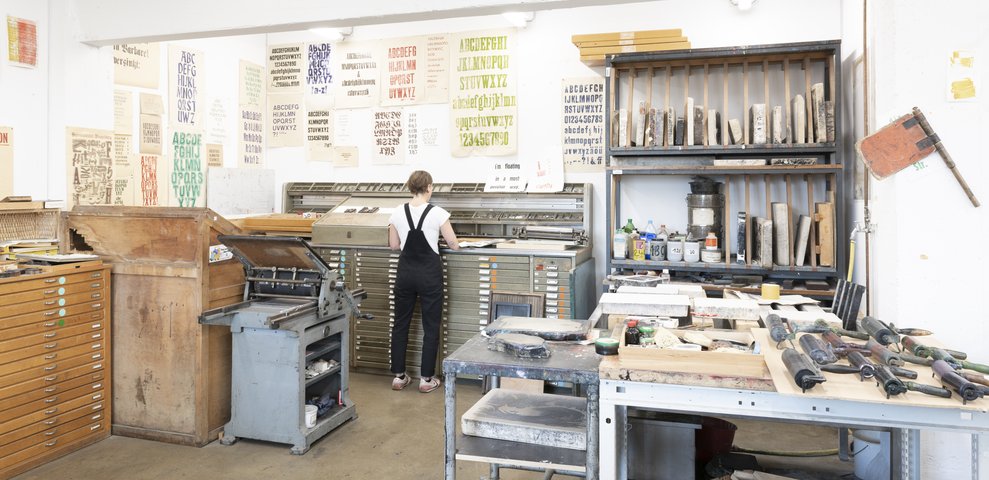 Workshop for Etching at HfK Bremen with a person working at a type cabinet, surrounded by tools, ink rollers, and various printed alphabets on the walls.