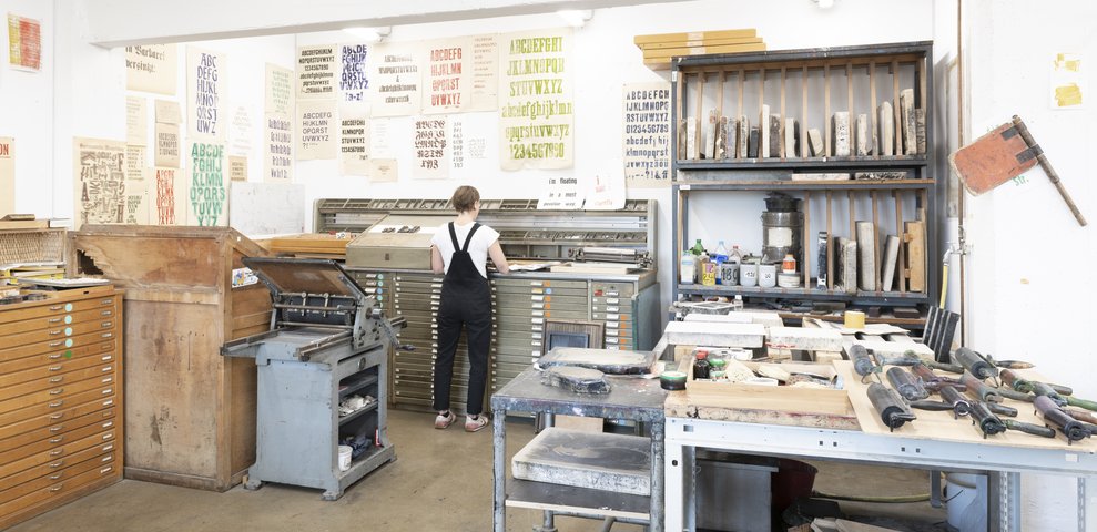 Workshop for Etching at HfK Bremen with a person working at a type cabinet, surrounded by tools, ink rollers, and various printed alphabets on the walls.