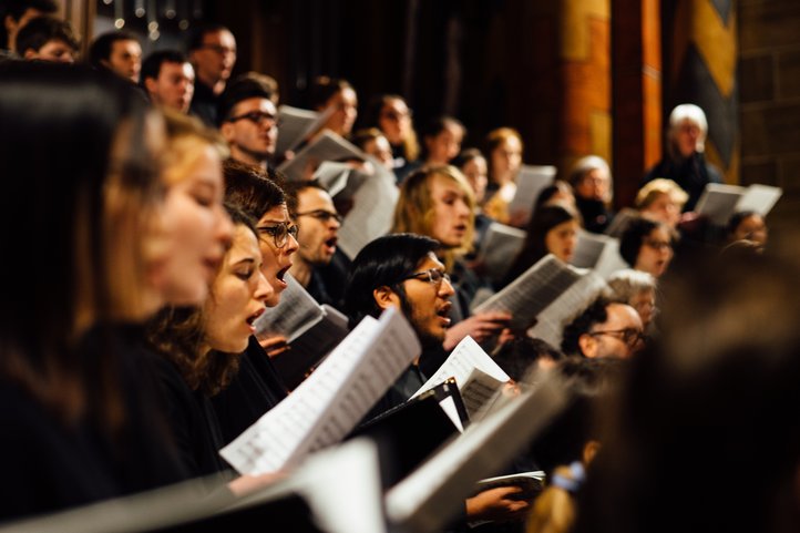 Choir singing in unison, holding sheet music in a dimly lit church.