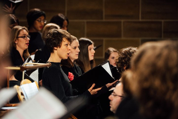 Chormitglieder singen leidenschaftlich, halten Notenblätter, vor einer Backsteinwand.