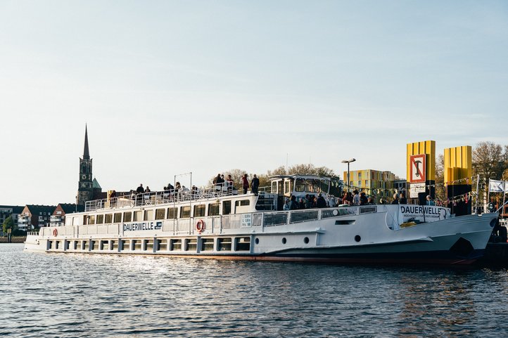 MS Dauerwelle am Liegeplatz, Seitenansicht mit Kirchturm und Stadtsilhouette.