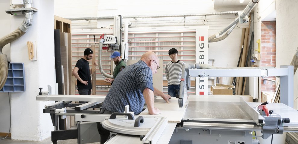 Werkstatt-Szene an der HfK Bremen: Personen arbeiten mit professioneller Ausstattung an Holzmodellen. In einer gut ausgestatteten Studio-Umgebung ist unter anderem eine große Tischsäge im Einsatz.