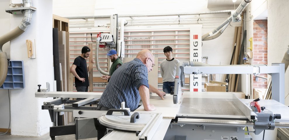 Werkstatt-Szene an der HfK Bremen: Personen arbeiten mit professioneller Ausstattung an Holzmodellen. In einer gut ausgestatteten Studio-Umgebung ist unter anderem eine große Tischsäge im Einsatz.