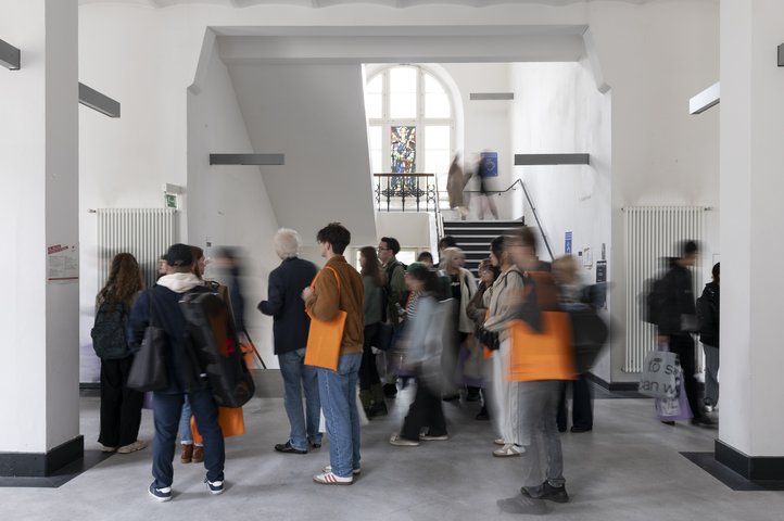 People in motion inside Dechanatstraße building, gathering and walking around with orange bags in a bright hallway.