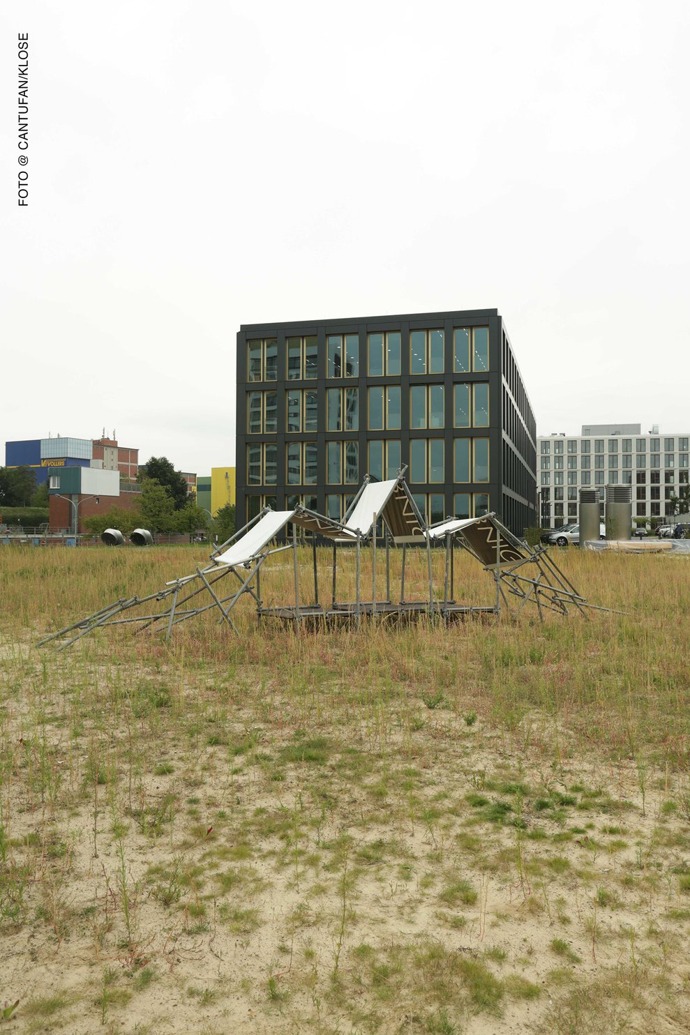 Skulpturale Konstruktion von Lucas Kalmus auf Brachfläche mit Gebäude im Hintergrund.