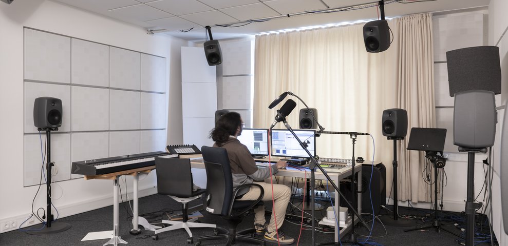 Audiolabor an der Hochschule für Künste Bremen mit einer Person, die an einem Computer mit Audio-Equipment arbeitet, umgeben von Lautsprechern, einem Keyboard und Mikrofonen.