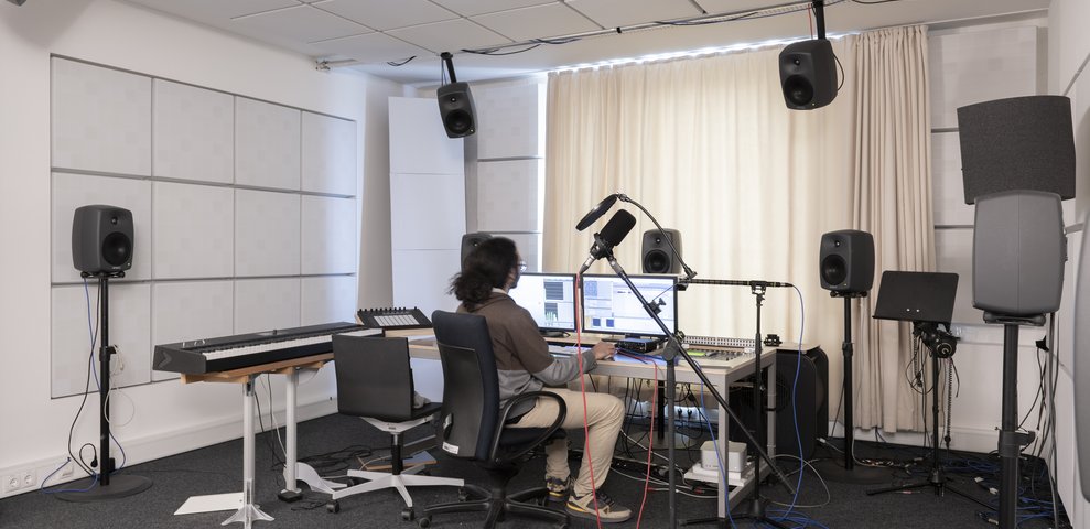Audiolab at HfK Bremen featuring a person working at a computer with audio equipment, surrounded by speakers, a keyboard, and microphones.