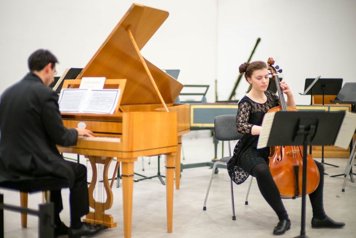 Musician playing a harpsichord with sheet music, and a cellist performing next to him, in a rehearsal setting