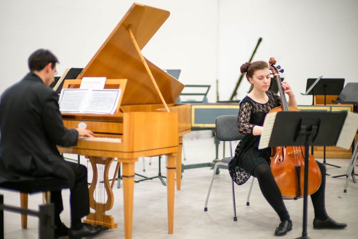Musician playing a harpsichord with sheet music, and a cellist performing next to him, in a rehearsal setting