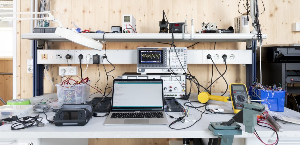 Electronics workstation at HfK Bremen featuring an oscilloscope, laptop displaying code, multimeter, wiring tools, and various electronic components.