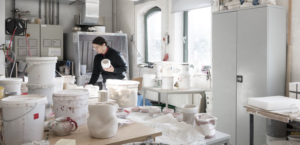Ceramics Workshop at HfK Bremen with an individual handling ceramics, surrounded by pots, tools, and shelves, well-lit by natural light.