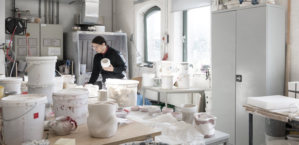 Keramikwerkstatt an der HfK Bremen mit einer Person, die Keramik bearbeitet, umgeben von Töpfen, Werkzeugen und Regalen, gut ausgeleuchtet durch Tageslicht.