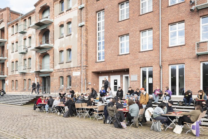 People gather and sit at tables outside Speicher XI of the HfK Bremen. The historic brick building features large windows and a stepped entrance, creating a welcoming atmosphere for students.
