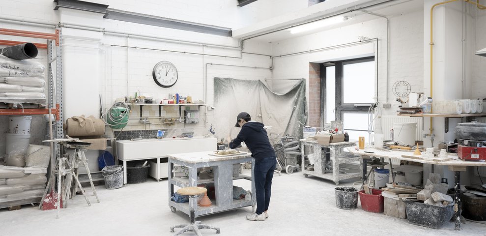 Eine Person arbeitet in der Gießerei der Hochschule für Künste Bremen, umgeben von Werkzeugen, Materialien und Geräten in einem geräumigen, gut beleuchteten Atelier.