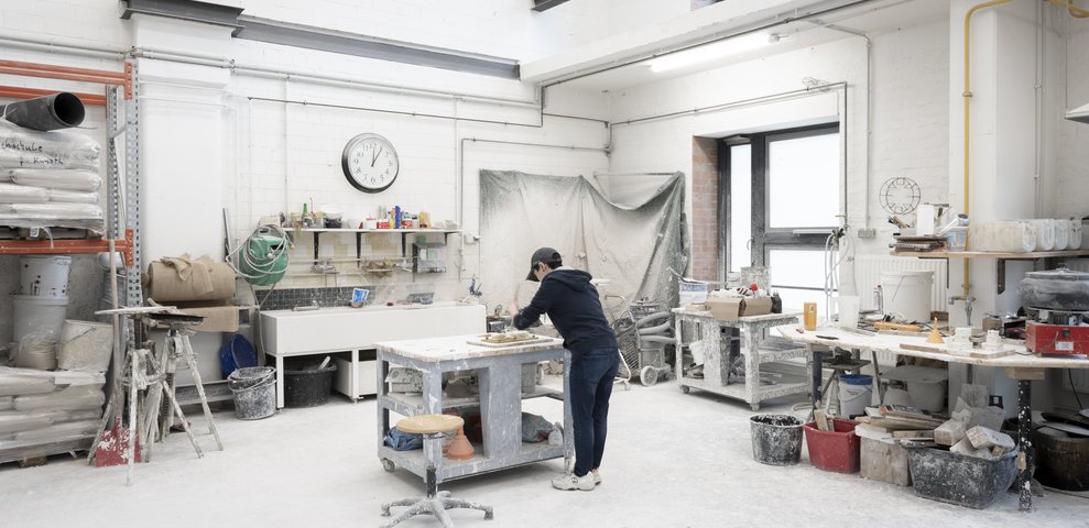 An individual works in the Foundry and Chasing Workshop at the University of the Arts Bremen, surrounded by tools, materials, and equipment in a spacious, well-lit studio.