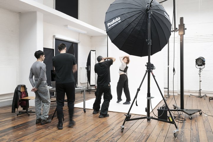 Photography studio session at HfK Bremen featuring a photographer capturing a model posing against a white backdrop. Studio lights and equipment are visible, with two onlookers observing the scene.